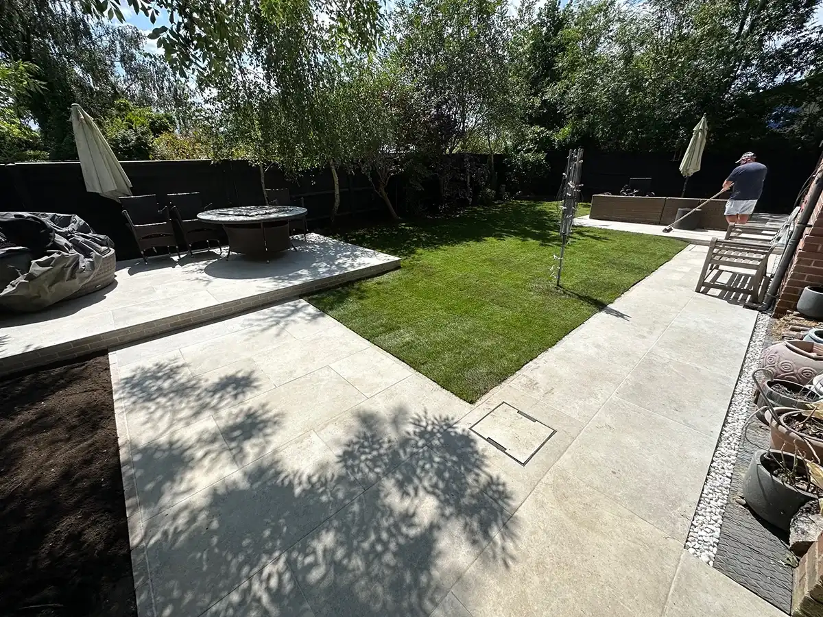 A well-maintained garden with a grassy area, patio, outdoor furniture, and a man using a broom under sunlight.