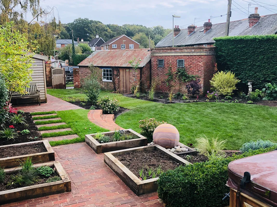 A landscaped garden featuring brick paths, wooden planters, a small shed, and a quaint brick building in the background.