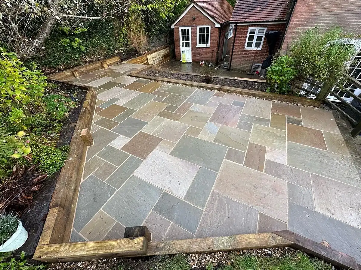 A newly paved outdoor area with irregular stone tiles in earthy tones, bordered by wooden planks, near a brick house and greenery.